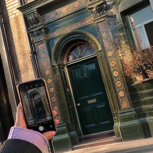 Charming ink and watercolor painting, clean lines, vibrant but soft color palette, storybook style. A close-up photo taken by a young woman's hand holding a phone camera. The subject is a beautiful old dark green tiled doorway on a London street, with ornate mosaic patterns around the frame and warm light hitting the tiles. The photo is slightly tilted as if taken casually while walking. No people visible. No readable text
Character consistency instruction: preserve these exact recurring appearance details: Maya: gender: Female; age: 25; skin tone: Light brown; hair color: Auburn; hair style: Shoulder-length braids; eye color: Warm brown; clothing: A lavender knitted sweater worn under a dark wool winter coat.; other features: Often seen holding a smartphone; has a cautious but charming half-smile. Do not add readable text unless the prompt explicitly asks for visible text.