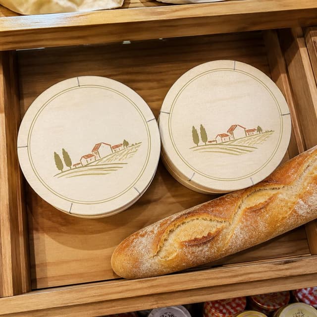 Charming ink and watercolor painting, clean lines, vibrant but soft color palette, storybook style. A top-down photo of two round camembert cheese boxes side by side on a wooden supermarket shelf, with a baguette resting alongside them. The boxes are cream-colored with simple circular lids. Warm, cheerful lighting. No readable text
Chat attachment instruction: Generate exactly one in-world chat attachment, not a collage, split-screen, comic panel, flashback, or explanatory scene. If the prompt is a screenshot, phone screen, document, note, card, map, receipt, label, object photo, or photo someone took, frame that artifact directly. A name appearing in visible text or in 'photo taken by X' is not an instruction to draw that person. Do not show recurring story participants as visible people unless the prompt itself explicitly says they are visible. Keep unrequested people, faces, and bodies out of the image.