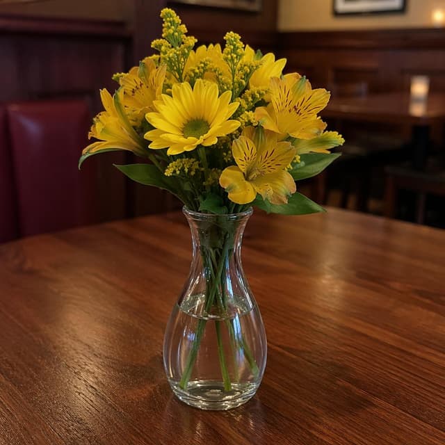 A single glass vase holding fresh yellow flowers, sitting on a plain bare wooden restaurant table
Chat attachment instruction: Generate exactly one in-world chat attachment, not a collage, split-screen, comic panel, flashback, or explanatory scene. If the prompt is a screenshot, phone screen, document, note, card, map, receipt, label, object photo, or photo someone took, frame that artifact directly. A name appearing in visible text or in 'photo taken by X' is not an instruction to draw that person. Do not show recurring story participants as visible people unless the prompt itself explicitly says they are visible. Keep unrequested people, faces, and bodies out of the image.