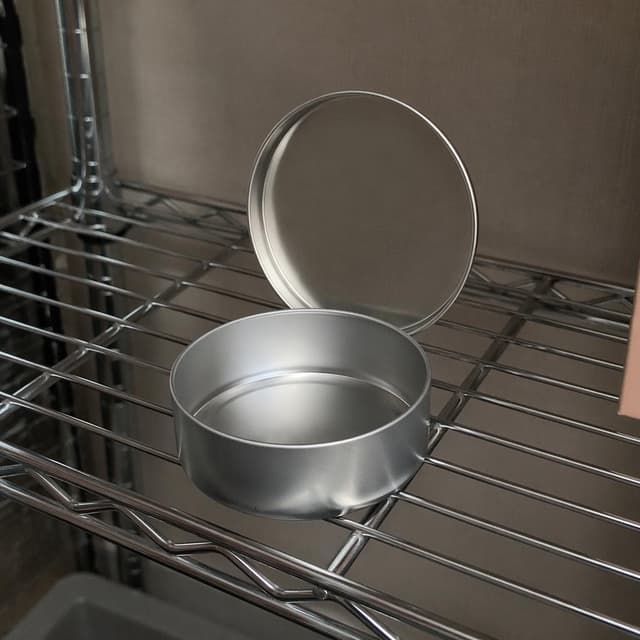 A small round metal tin with its lid open, completely empty, resting on a metal shelf
Chat attachment instruction: Generate exactly one in-world chat attachment, not a collage, split-screen, comic panel, flashback, or explanatory scene. If the prompt is a screenshot, phone screen, document, note, card, map, receipt, label, object photo, or photo someone took, frame that artifact directly. A name appearing in visible text or in 'photo taken by X' is not an instruction to draw that person. Do not show recurring story participants as visible people unless the prompt itself explicitly says they are visible. Keep unrequested people, faces, and bodies out of the image.