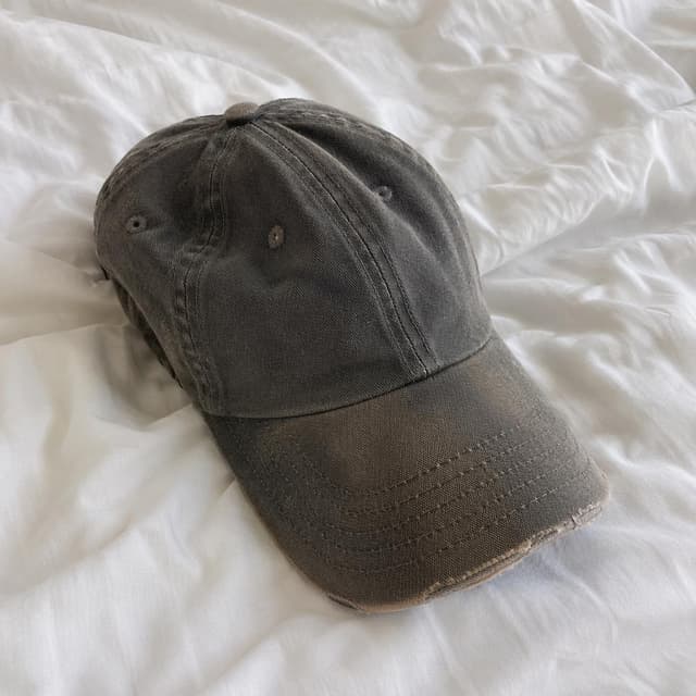a single worn-out, faded baseball cap resting on plain white bed sheets
Chat attachment instruction: Generate exactly one in-world chat attachment, not a collage, split-screen, comic panel, flashback, or explanatory scene. If the prompt is a screenshot, phone screen, document, note, card, map, receipt, label, object photo, or photo someone took, frame that artifact directly. A name appearing in visible text or in 'photo taken by X' is not an instruction to draw that person. Do not show recurring story participants as visible people unless the prompt itself explicitly says they are visible. Keep unrequested people, faces, and bodies out of the image.