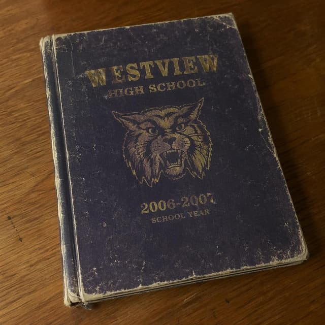 A worn-out school yearbook from 2007, lying on a wooden desk, showing a faded cover with the school name
Chat attachment instruction: Generate exactly one in-world chat attachment, not a collage, split-screen, comic panel, flashback, or explanatory scene. If the prompt is a screenshot, phone screen, document, note, card, map, receipt, label, object photo, or photo someone took, frame that artifact directly. A name appearing in visible text or in 'photo taken by X' is not an instruction to draw that person. Do not show recurring story participants as visible people unless the prompt itself explicitly says they are visible. Keep unrequested people, faces, and bodies out of the image. Do not add readable text unless the prompt explicitly asks for visible text.
