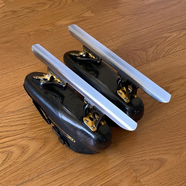 A pair of short-track speed skates with long silver blades, resting blade-side up on a plain wooden floor
Chat attachment instruction: Generate exactly one in-world chat attachment, not a collage, split-screen, comic panel, flashback, or explanatory scene. If the prompt is a screenshot, phone screen, document, note, card, map, receipt, label, object photo, or photo someone took, frame that artifact directly. A name appearing in visible text or in 'photo taken by X' is not an instruction to draw that person. Do not show recurring story participants as visible people unless the prompt itself explicitly says they are visible. Keep unrequested people, faces, and bodies out of the image.