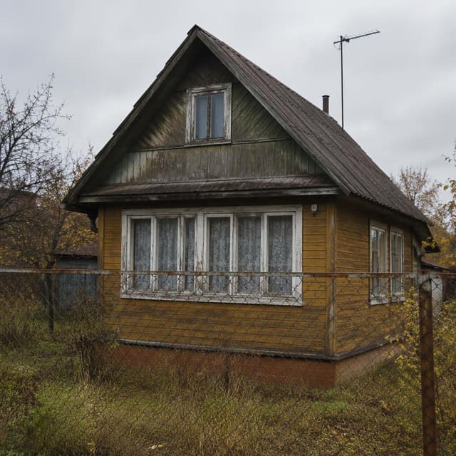 A small, weathered wooden dacha cottage seen from outside a wire fence, plain overcast sky behind it, no people
Chat attachment instruction: Generate exactly one in-world chat attachment, not a collage, split-screen, comic panel, flashback, or explanatory scene. If the prompt is a screenshot, phone screen, document, note, card, map, receipt, label, object photo, or photo someone took, frame that artifact directly. A name appearing in visible text or in 'photo taken by X' is not an instruction to draw that person. Do not show recurring story participants as visible people unless the prompt itself explicitly says they are visible. Keep unrequested people, faces, and bodies out of the image.