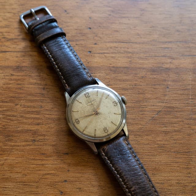 an old wristwatch on a plain wooden table
Chat attachment instruction: Generate exactly one in-world chat attachment, not a collage, split-screen, comic panel, flashback, or explanatory scene. If the prompt is a screenshot, phone screen, document, note, card, map, receipt, label, object photo, or photo someone took, frame that artifact directly. A name appearing in visible text or in 'photo taken by X' is not an instruction to draw that person. Do not show recurring story participants as visible people unless the prompt itself explicitly says they are visible. Keep unrequested people, faces, and bodies out of the image.