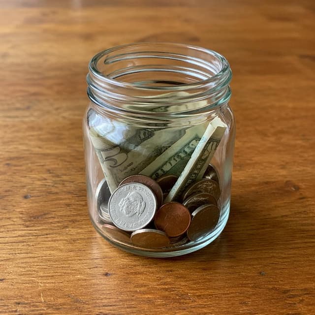 a small glass jar with coins and a few folded banknotes on a plain wooden table
Chat attachment instruction: Generate exactly one in-world chat attachment, not a collage, split-screen, comic panel, flashback, or explanatory scene. If the prompt is a screenshot, phone screen, document, note, card, map, receipt, label, object photo, or photo someone took, frame that artifact directly. A name appearing in visible text or in 'photo taken by X' is not an instruction to draw that person. Do not show recurring story participants as visible people unless the prompt itself explicitly says they are visible. Keep unrequested people, faces, and bodies out of the image.
