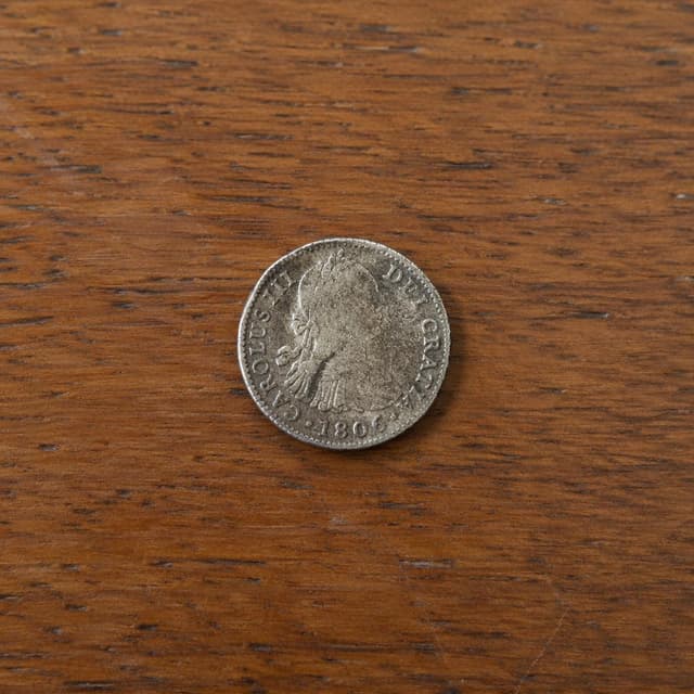 a small old silver coin on a plain wooden surface
Chat attachment instruction: Generate exactly one in-world chat attachment, not a collage, split-screen, comic panel, flashback, or explanatory scene. If the prompt is a screenshot, phone screen, document, note, card, map, receipt, label, object photo, or photo someone took, frame that artifact directly. A name appearing in visible text or in 'photo taken by X' is not an instruction to draw that person. Do not show recurring story participants as visible people unless the prompt itself explicitly says they are visible. Keep unrequested people, faces, and bodies out of the image.