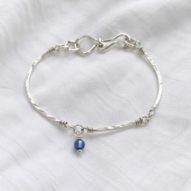 A handmade silver bracelet with a small blue bead charm, photographed on a plain white cloth background
Chat attachment instruction: Generate exactly one in-world chat attachment, not a collage, split-screen, comic panel, flashback, or explanatory scene. If the prompt is a screenshot, phone screen, document, note, card, map, receipt, label, object photo, or photo someone took, frame that artifact directly. A name appearing in visible text or in 'photo taken by X' is not an instruction to draw that person. Do not show recurring story participants as visible people unless the prompt itself explicitly says they are visible. Keep unrequested people, faces, and bodies out of the image.