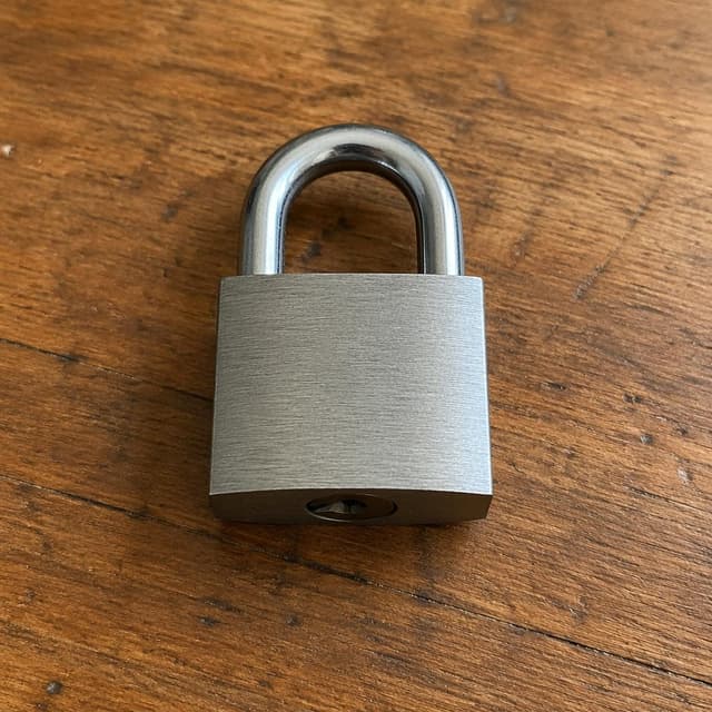 a plain metal padlock on a wooden table
Chat attachment instruction: Generate exactly one in-world chat attachment, not a collage, split-screen, comic panel, flashback, or explanatory scene. If the prompt is a screenshot, phone screen, document, note, card, map, receipt, label, object photo, or photo someone took, frame that artifact directly. A name appearing in visible text or in 'photo taken by X' is not an instruction to draw that person. Do not show recurring story participants as visible people unless the prompt itself explicitly says they are visible. Keep unrequested people, faces, and bodies out of the image.