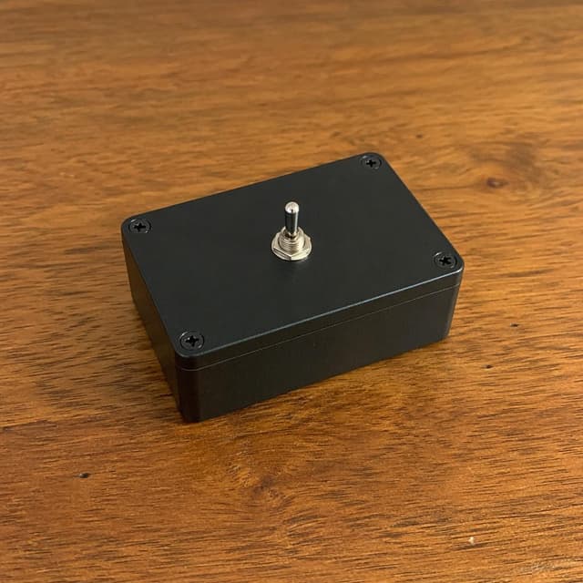 A small black electronic device with a single toggle switch on a plain wooden table
Chat attachment instruction: Generate exactly one in-world chat attachment, not a collage, split-screen, comic panel, flashback, or explanatory scene. If the prompt is a screenshot, phone screen, document, note, card, map, receipt, label, object photo, or photo someone took, frame that artifact directly. A name appearing in visible text or in 'photo taken by X' is not an instruction to draw that person. Do not show recurring story participants as visible people unless the prompt itself explicitly says they are visible. Keep unrequested people, faces, and bodies out of the image.