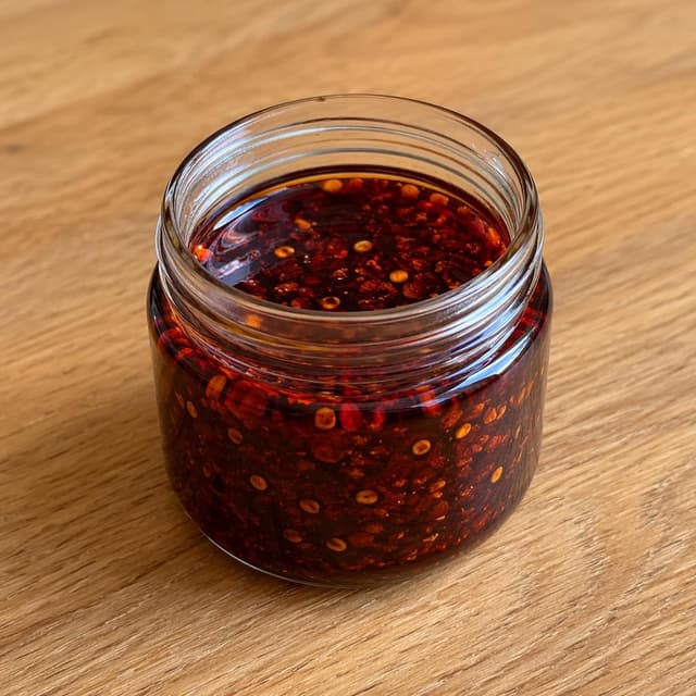 a small glass jar filled with dark red, glossy chili oil, sitting on a plain wooden table
Chat attachment instruction: Generate exactly one in-world chat attachment, not a collage, split-screen, comic panel, flashback, or explanatory scene. If the prompt is a screenshot, phone screen, document, note, card, map, receipt, label, object photo, or photo someone took, frame that artifact directly. A name appearing in visible text or in 'photo taken by X' is not an instruction to draw that person. Do not show recurring story participants as visible people unless the prompt itself explicitly says they are visible. Keep unrequested people, faces, and bodies out of the image.