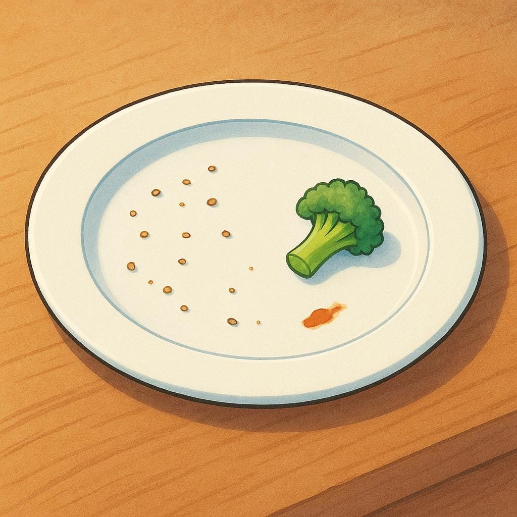 A white ceramic dinner plate sitting on a table, holding a few small scraps of uneaten food, like crumbs and a few vegetable pieces.