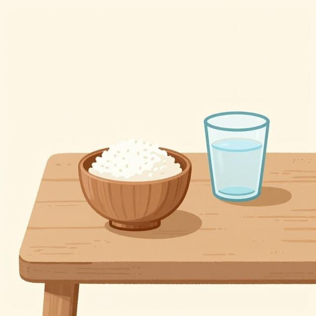 A small bowl containing a few grains of rice and a single glass of water on a plain wooden table.